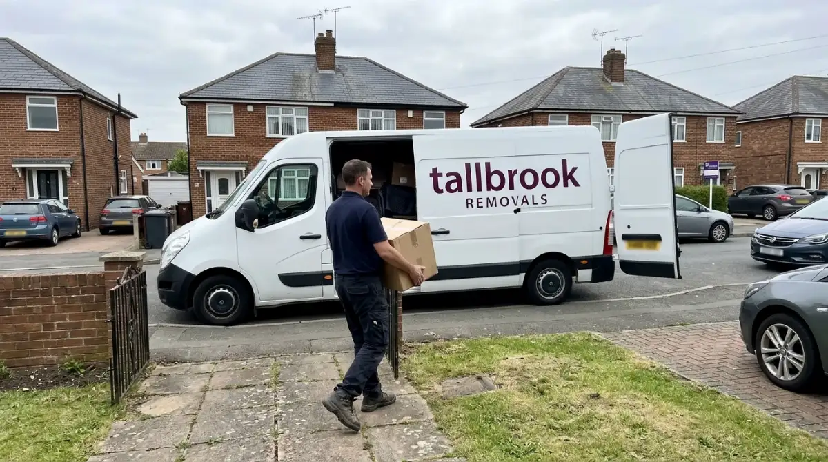 Mover carrying a cardboard box across a front garden towards a white Tallbrook Removals van
